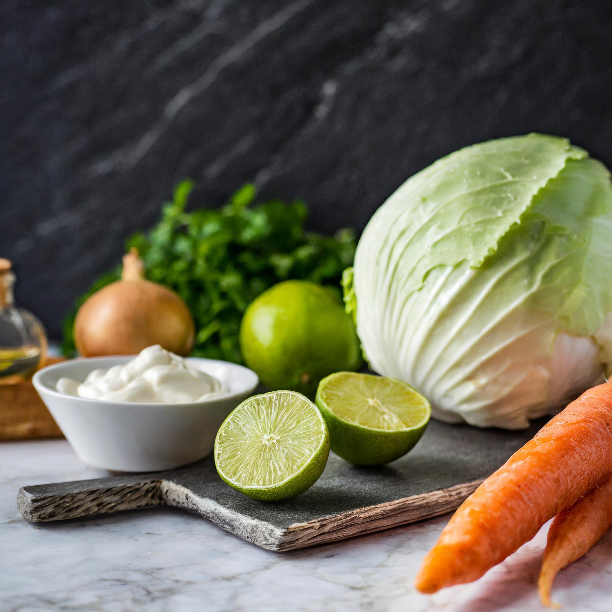 Frische Zutaten auf einer rustikalen, weiß gefliesten Oberfläche: ein ganzer Kohlkopf, zwei Karotten, eine halbierte Limette, eine Schale mit Sauerrahm, eine Zwiebel und ein Bund Koriander. Im Hintergrund ist eine Flasche Olivenöl und eine weitere Limette zu sehen, alles vor einer schwarzen, strukturierten Schieferwand.