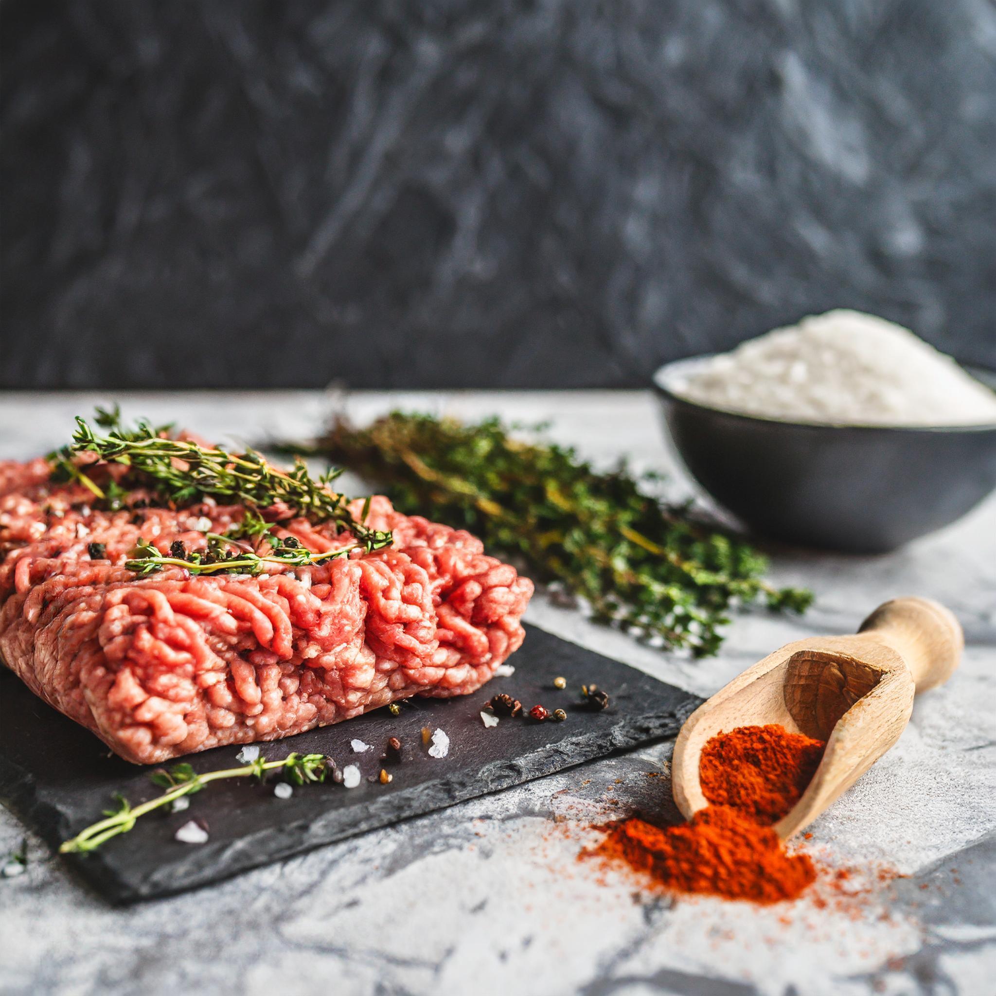 Ein rohes Stück Hackfleisch, dekoriert mit frischen Thymianzweigen und Meersalz, liegt auf einer schwarzen Schieferplatte. Im Vordergrund sind gemahlener Paprika in einem Holzlöffel und ein paar Pfefferkörner zu sehen. Im Hintergrund stehen eine Schale mit Meersalz und ein Bund frischer Kräuter, alles arrangiert auf einer rustikalen, weiß gefliesten Oberfläche vor einer schwarzen, strukturierten Schieferwand.