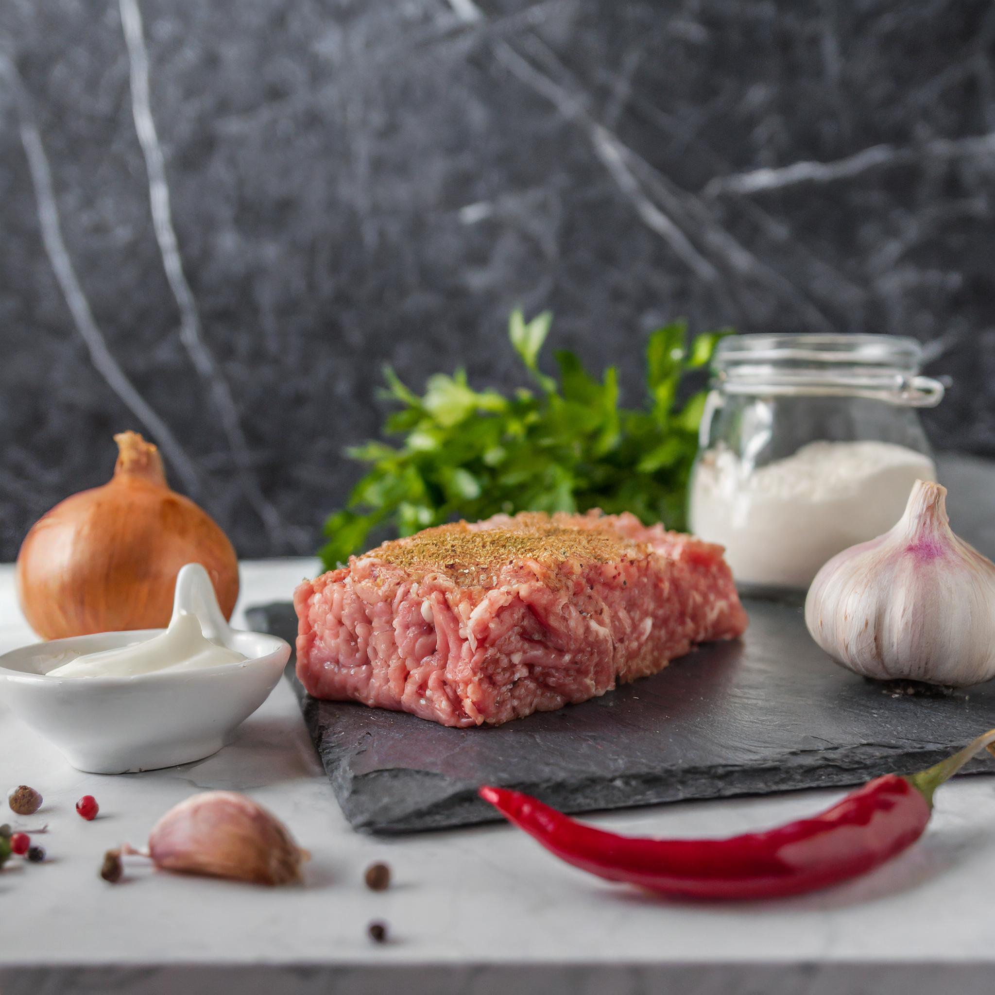 Ein Arrangement von Zutaten auf einer Schieferplatte: ein Stück rohes Hackfleisch mit Gewürzen bestreut, eine Zwiebel, eine Knoblauchknolle, eine rote Chilischote, eine Schale mit Joghurt, ein Glas Mehl und frische Petersilie, vor einem dunklen, marmorierten Hintergrund.