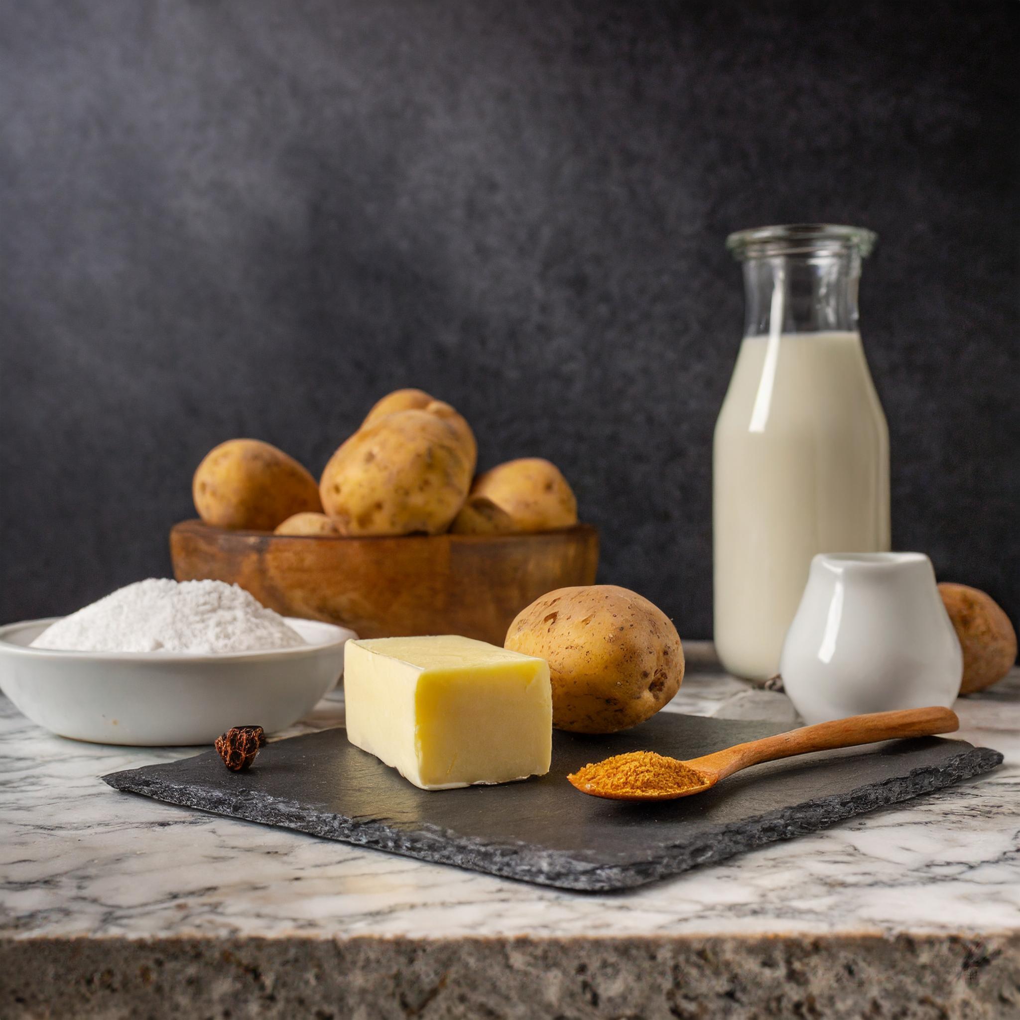 Eine Auswahl an Zutaten für ein Rezept, darunter rohe Kartoffeln in einer Holzschale, ein Stück Butter auf einer Schieferplatte, eine Schüssel mit Mehl, eine Flasche Milch, eine kleine Kanne und ein Holzlöffel mit Gewürzpulver, arrangiert auf einer Marmortischplatte vor einem dunklen, strukturierten Hintergrund.