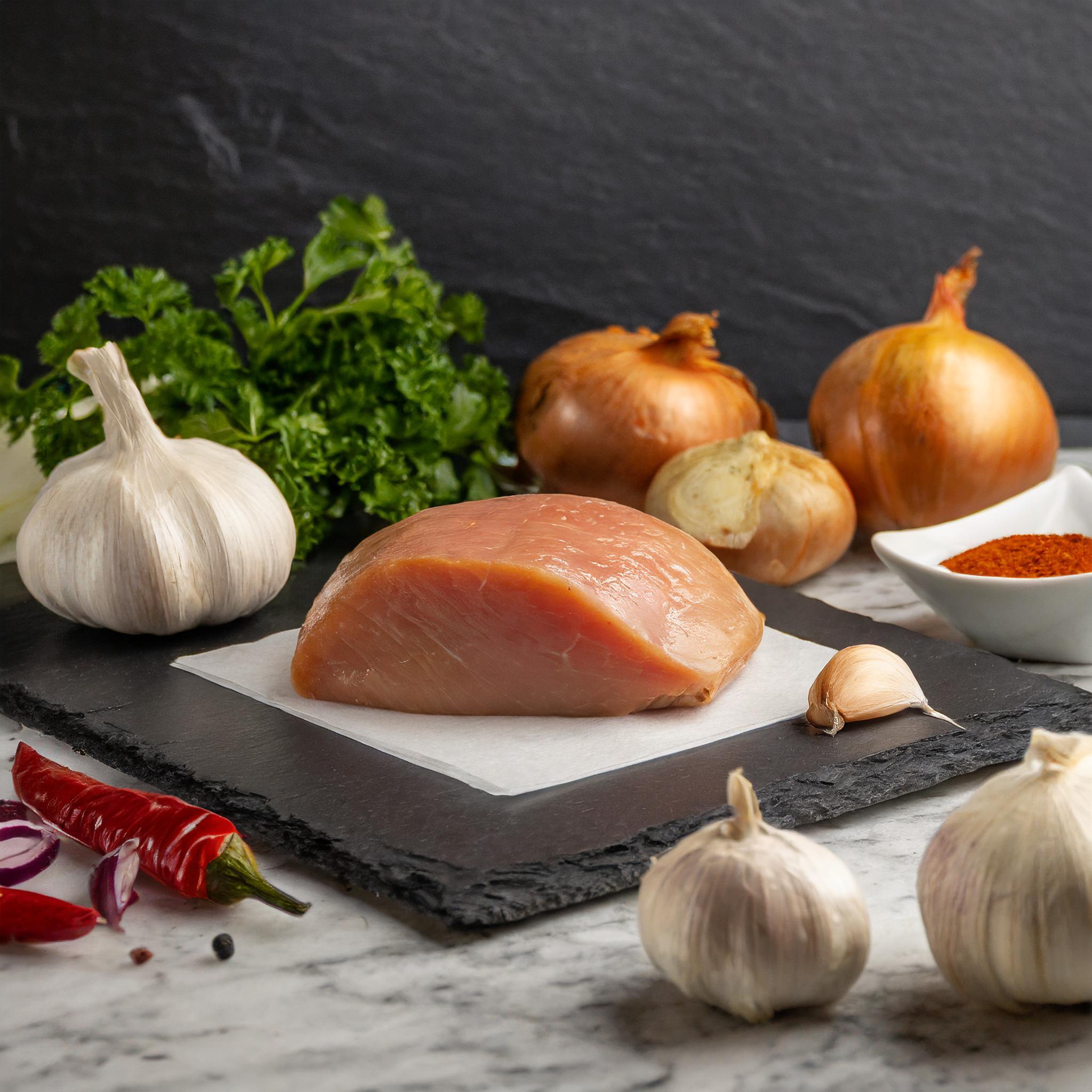Ein Arrangement von Zutaten auf einer Schieferplatte: ein Stück rohes Hähnchenbrustfilet, Knoblauchknollen, Zwiebeln, frische Petersilie, rote Chilischoten und eine Schale mit Paprikapulver, arrangiert vor einem dunklen, strukturierten Hintergrund.