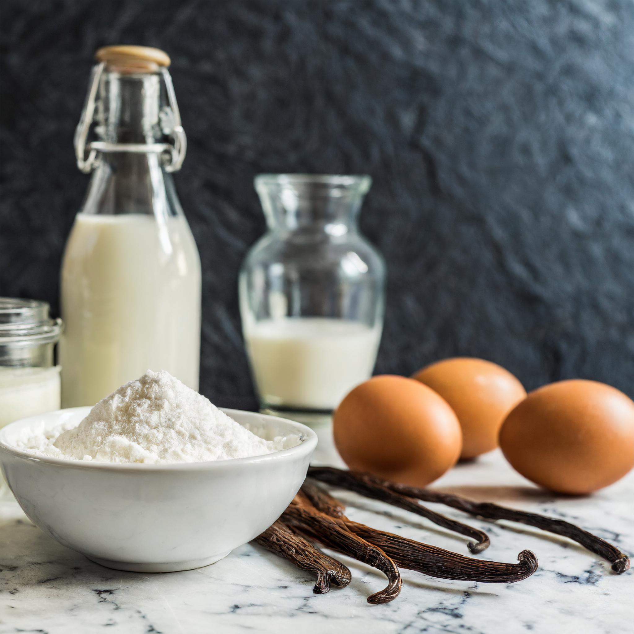 Nahaufnahme eines rustikalen weißen Granittischs vor einem schwarzen, strukturierten Schieferhintergrund, auf dem ein Glas Vanillepudding dekorativ arrangiert ist.