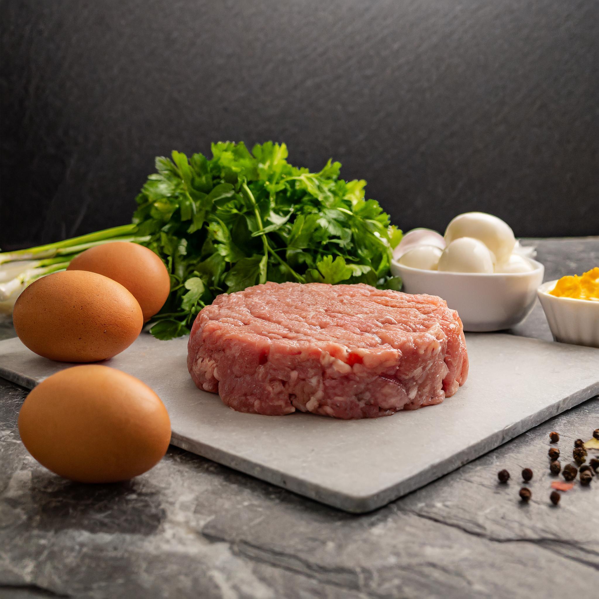Ein Arrangement von Zutaten auf einer Schieferplatte: ein roher Fleischpattie, drei Eier, frische Petersilie, eine Schüssel mit gekochten Eiern und eine Schüssel mit Senf. Im Hintergrund sind Frühlingszwiebeln zu sehen, alles arrangiert vor einem dunklen, strukturierten Hintergrund.