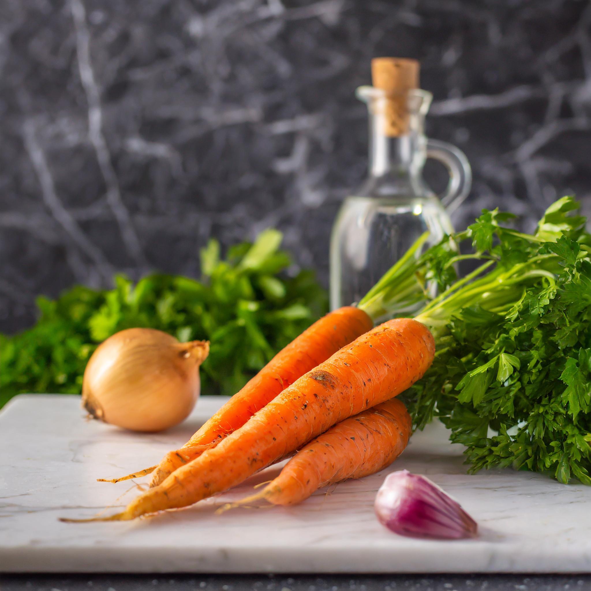 Ein Arrangement von frischen Zutaten auf einer Marmoroberfläche: mehrere Karotten mit Grün, eine Zwiebel, eine Knoblauchzehe, frische Petersilie und eine Flasche Olivenöl vor einem dunklen, marmorierten Hintergrund.