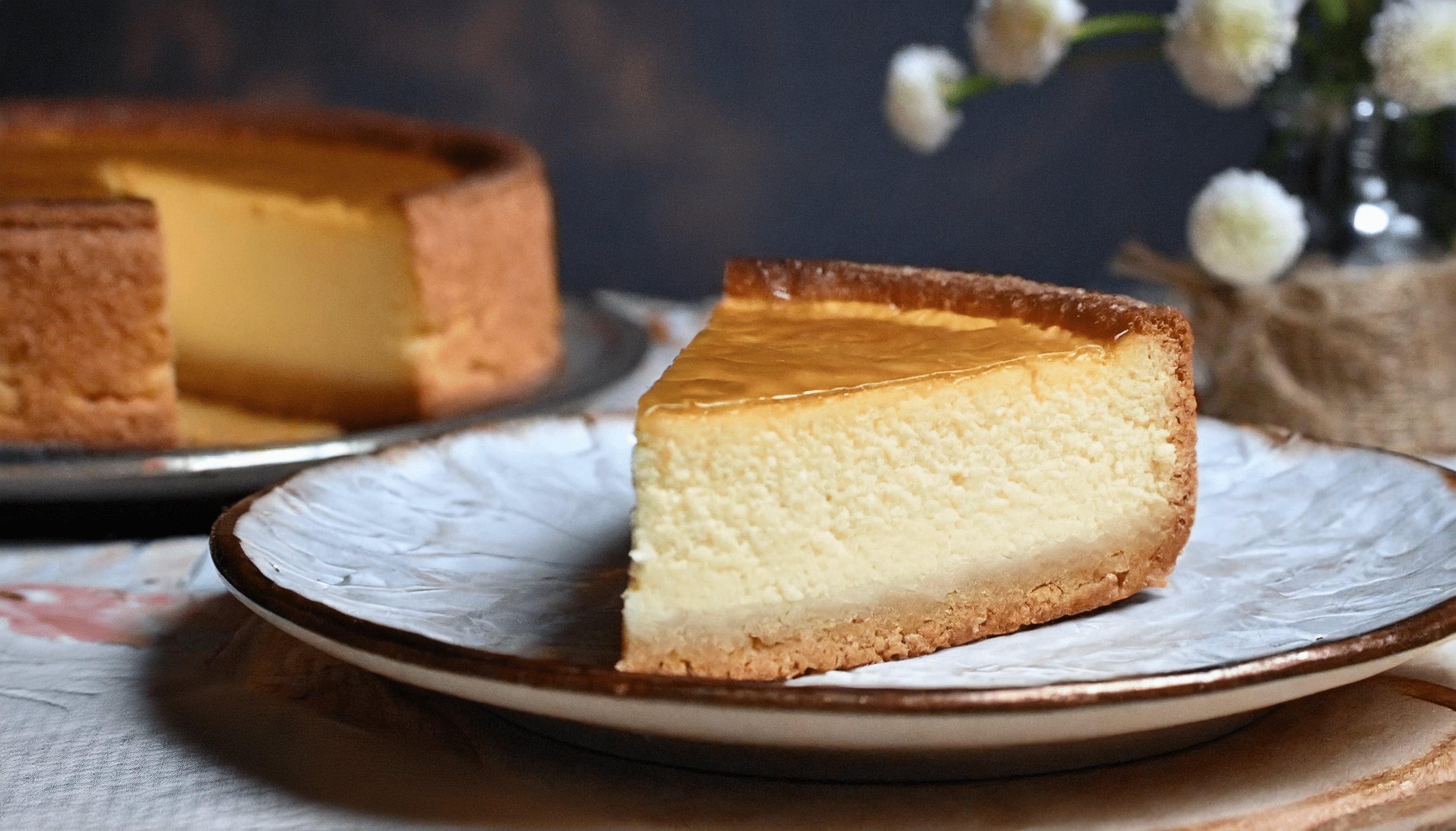 Ein Stück cremiger Käsekuchen mit goldbrauner Kruste auf einem rustikalen Teller. Im Hintergrund ist der restliche Kuchen auf einer Kuchenplatte zu sehen, dekoriert mit weißen Blumen in einer Vase. Alles ist vor einem dunklen, strukturierten Hintergrund arrangiert.