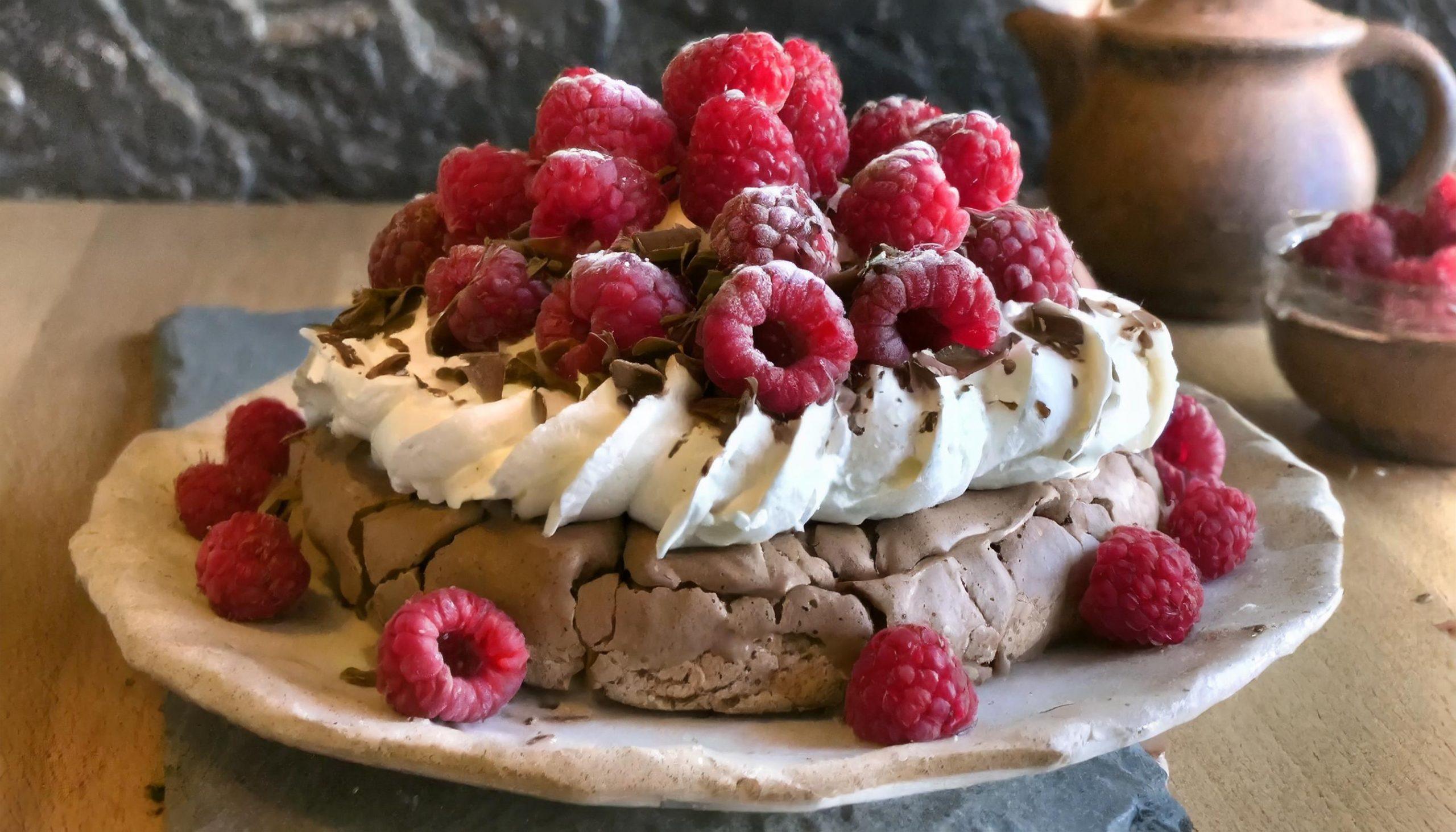 Nahaufnahme eines rustikalen weißen Granittischs vor einem schwarzen, strukturierten Schieferhintergrund, auf dem eine dekorativ arrangierte Pavlova mit frischen Beeren steht.