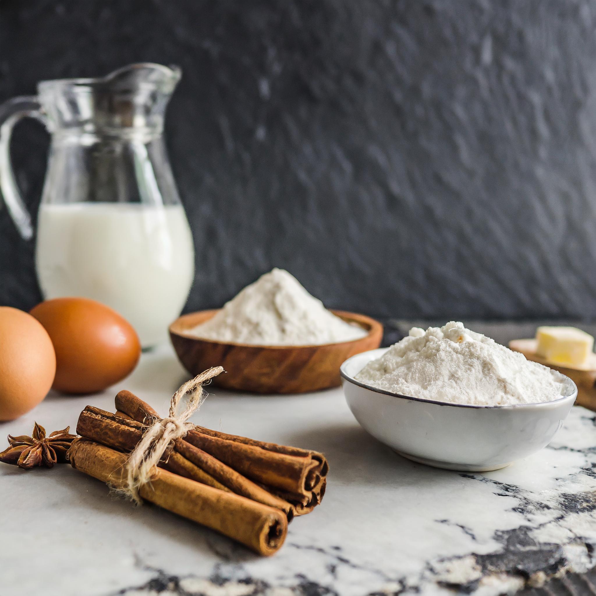Frische Backzutaten auf einer weißen Marmorplatte: zwei braune Eier, mehrere Zimtstangen, und Anisstern liegen neben Schalen mit Mehl. Im Hintergrund steht eine Glaskaraffe mit Milch, alles arrangiert vor einer schwarzen, strukturierten Schieferwand.