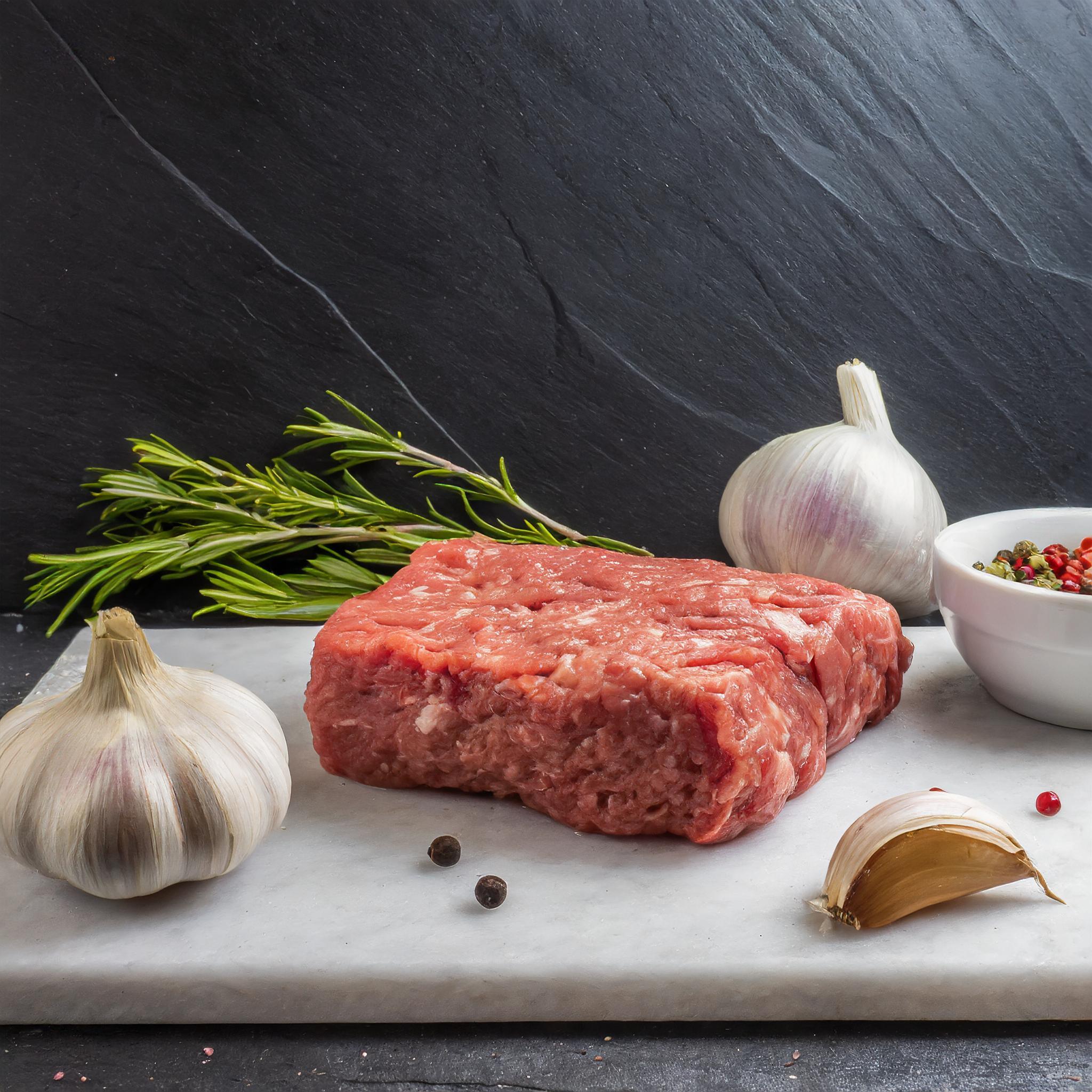Ein rohes Stück Hackfleisch auf einer weißen Marmorplatte, flankiert von zwei Knoblauchknollen und einem Zweig Rosmarin. Daneben steht eine kleine weiße Schale mit buntem Pfeffer. Im Hintergrund sieht man eine schwarze, strukturierte Schieferwand.