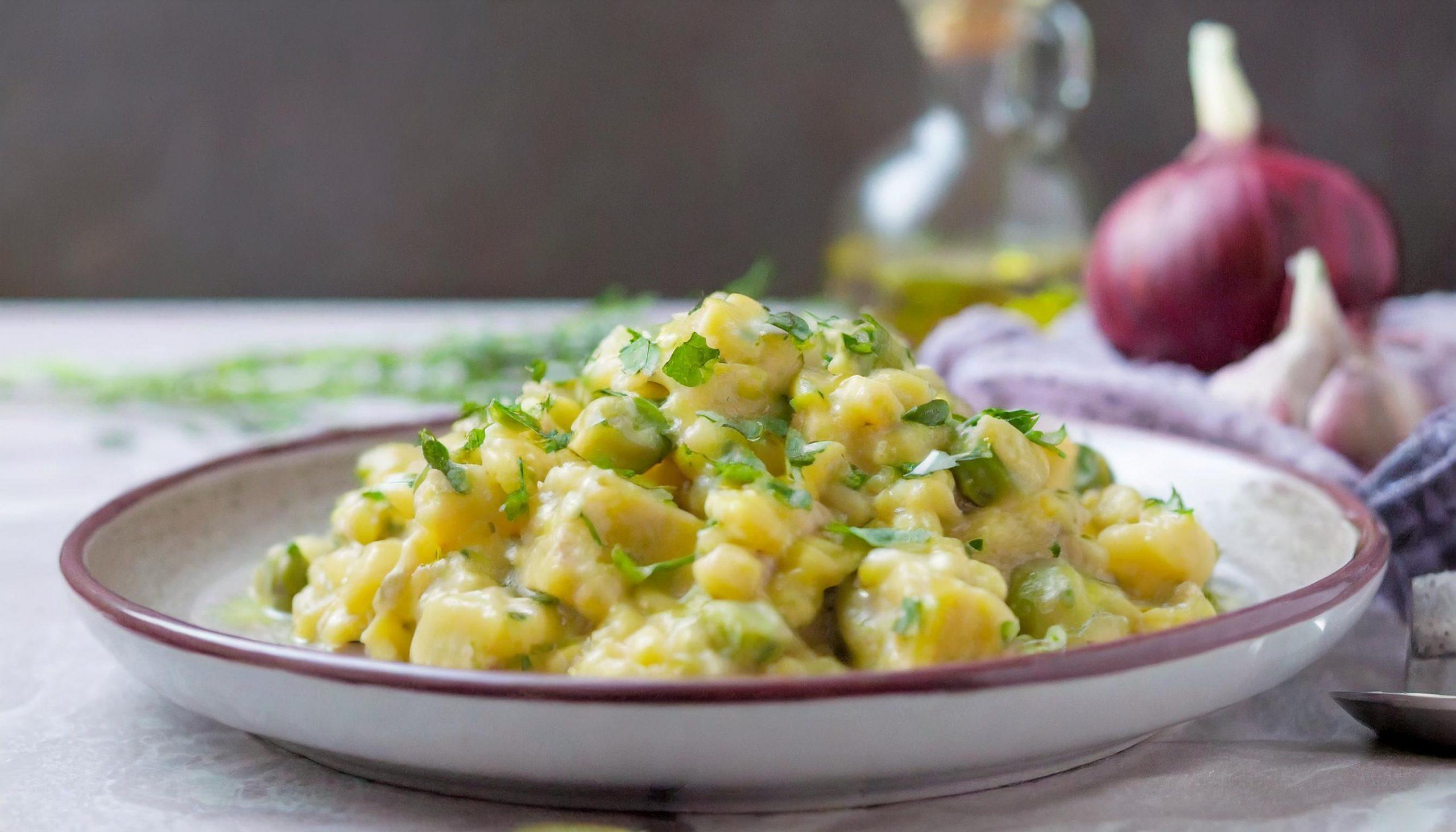 Nahaufnahme eines rustikalen weißen Granittischs vor einem schwarzen, strukturierten Schieferhintergrund, auf dem eine Schüssel mit traditionellem Kartoffelsalat dekorativ arrangiert ist.