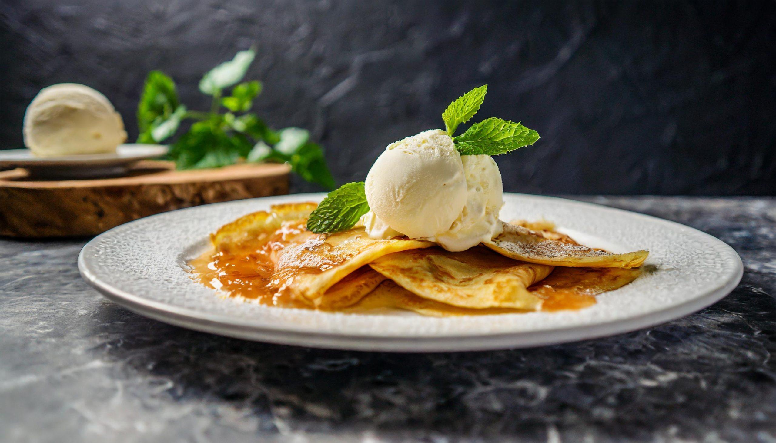 Nahaufnahme eines rustikalen weißen Granittischs vor einem schwarzen, strukturierten Schieferhintergrund, auf dem ein Teller mit Crêpe Suzette, garniert mit Orangenscheiben und Sauce, dekorativ arrangiert ist.