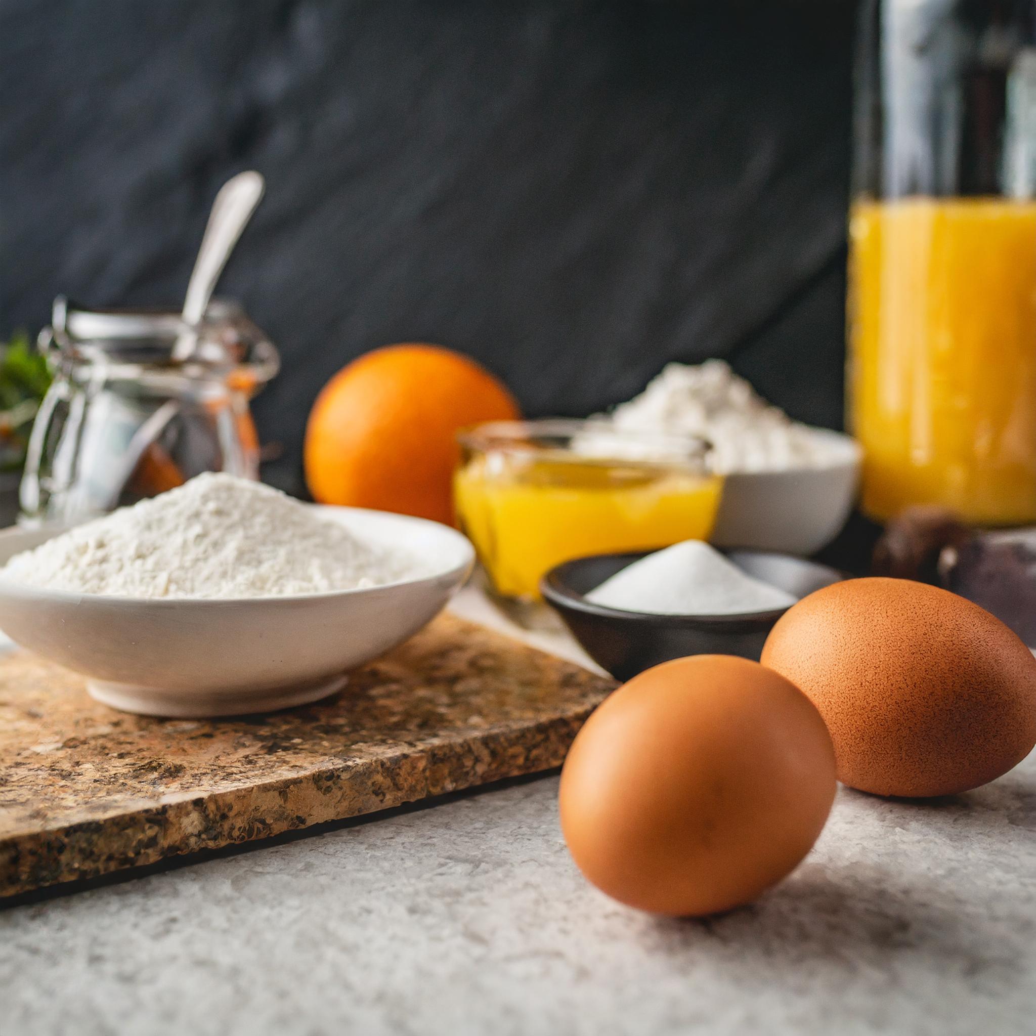 Eine Nahaufnahme eines rustikalen weißen Granittisches, auf dem verschiedene Zutaten für das Backen arrangiert sind. Auf dem Tisch liegen zwei braune Eier, eine Schale mit Mehl, eine Schale mit Zucker, eine Schale mit Butter, ein Glas mit Orangensaft, eine Orange und eine Flasche mit Vanilleextrakt. Im Hintergrund sind ein Glas mit Honig und eine weitere Orange zu sehen. Der Hintergrund besteht aus schwarzem, strukturiertem Schiefer.