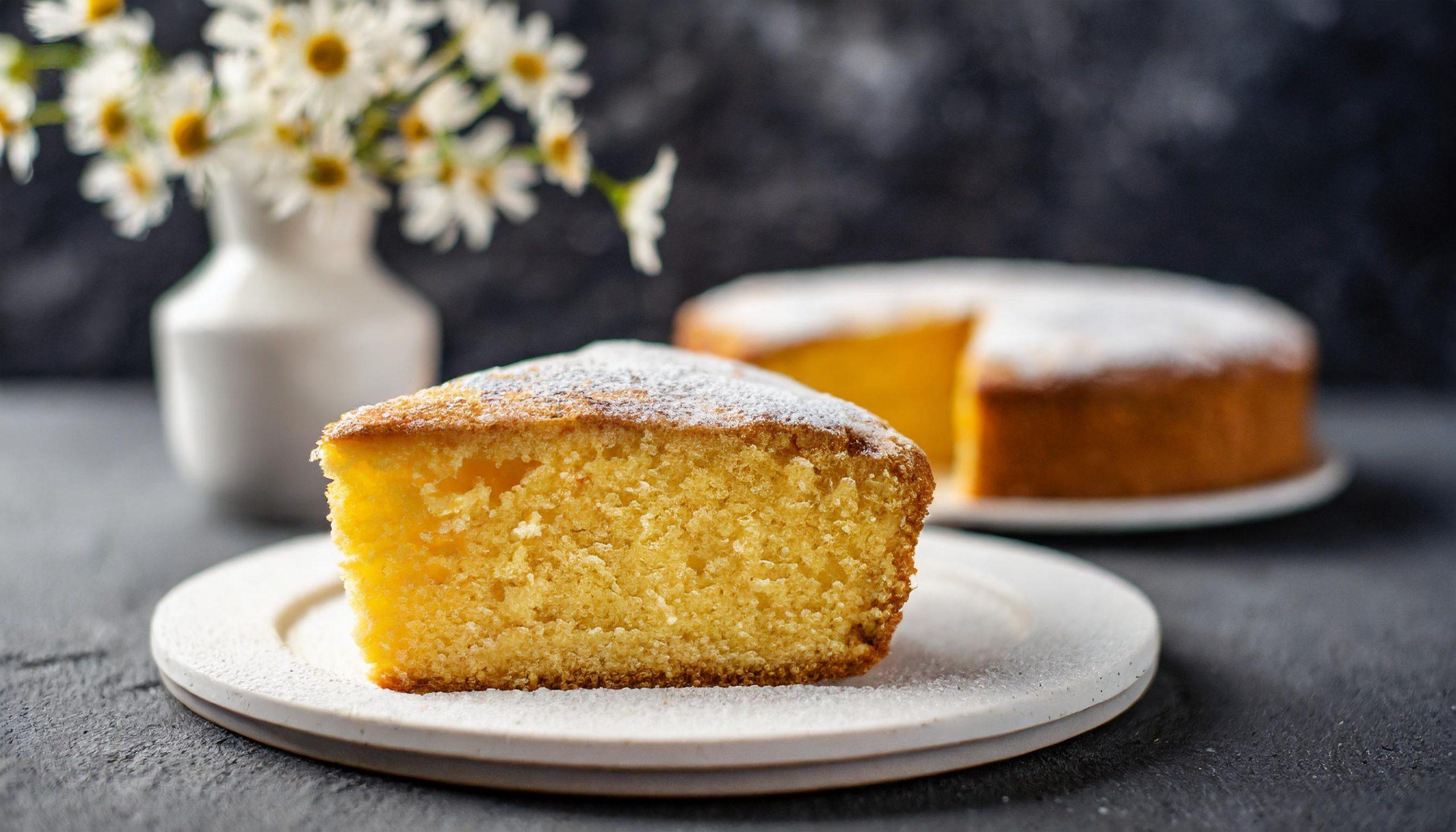Ein saftiges Stück Zitronenkuchen, bestäubt mit Puderzucker, liegt auf einem weißen Teller vor einem dunklen, strukturierten Hintergrund. Im Hintergrund ist der restliche Kuchen auf einem weiteren Teller zu sehen. Eine weiße Vase mit weißen Blumen ergänzt die rustikale Szene.