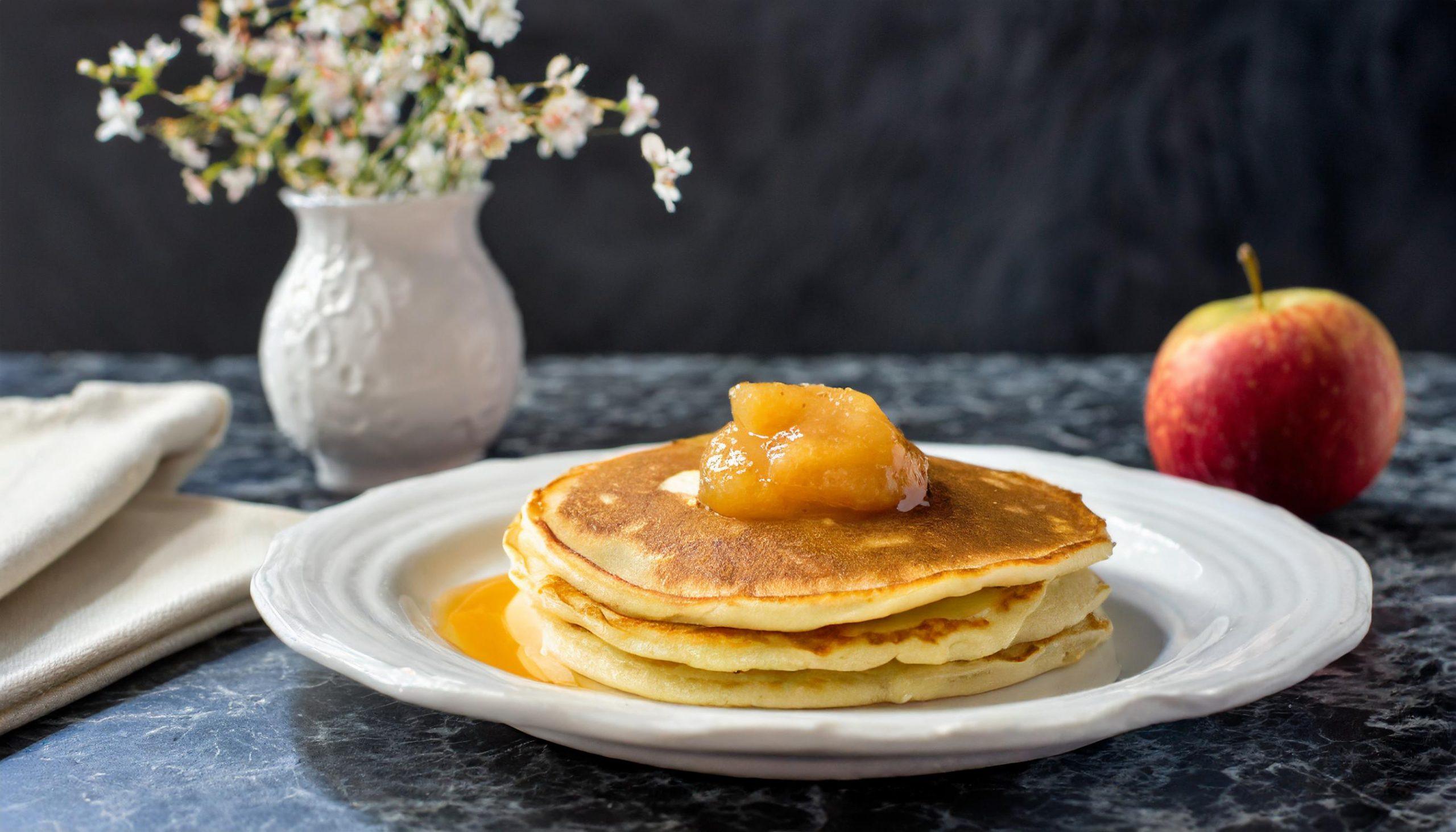 Ein Stapel goldbrauner Pfannkuchen, garniert mit einem Klecks Apfelmus, auf einem weißen Teller. Im Hintergrund steht eine weiße Vase mit zarten Blumen und ein roter Apfel auf einer rustikalen weißen Granitplatte vor einem dunkelgrauen, strukturierten Schieferhintergrund. Eine beiger Stoffserviette liegt links daneben.