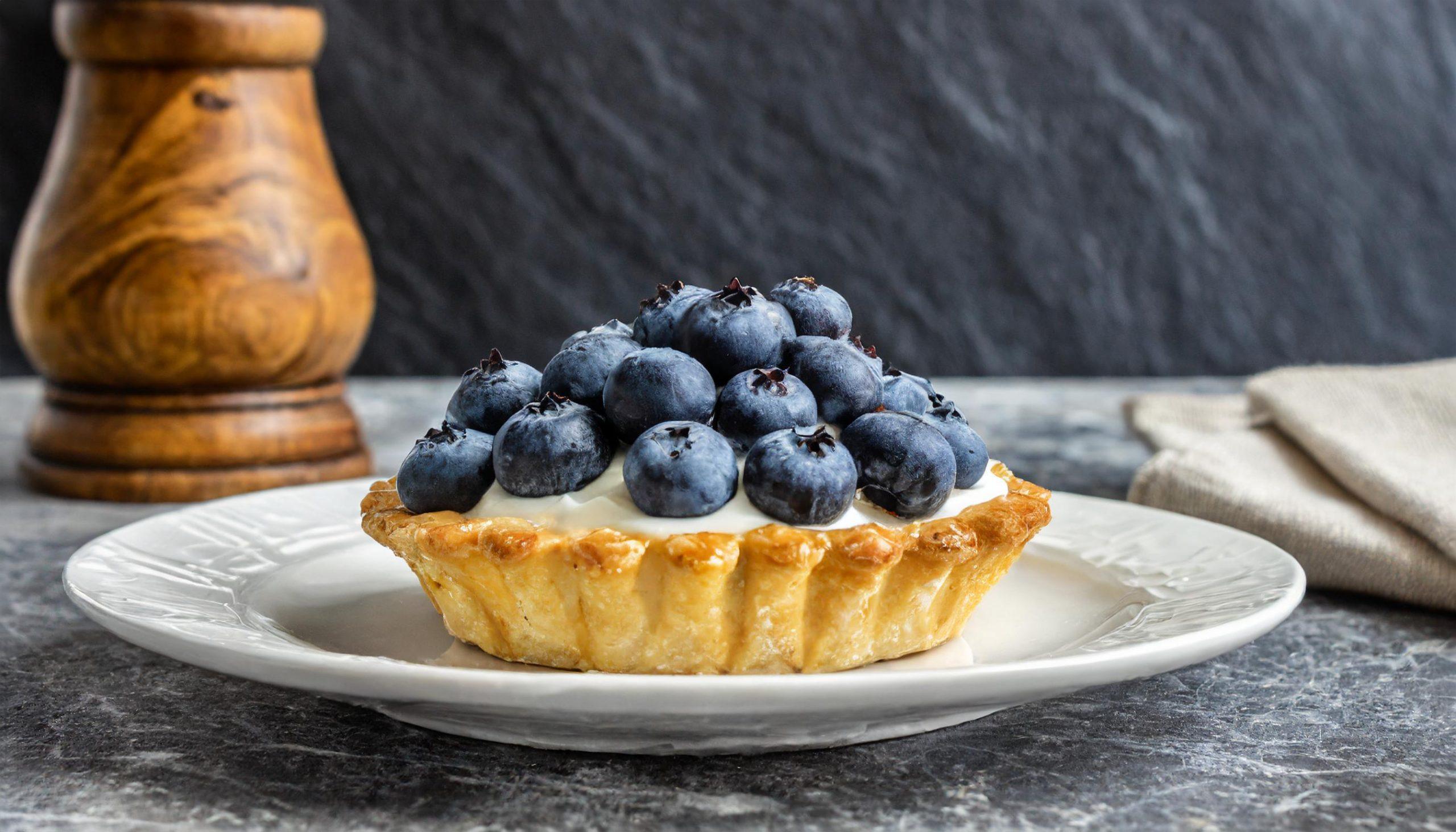 Ein köstliches Blaubeertörtchen auf einem weißen Teller, gefüllt mit cremiger Füllung und bedeckt mit frischen Blaubeeren. Im Hintergrund ist ein hölzerner Pfefferstreuer und ein beiger Stoffserviette zu sehen. Die Szene spielt sich auf einer rustikalen weißen Granitplatte vor einem dunkelgrauen, strukturierten Schieferhintergrund ab.