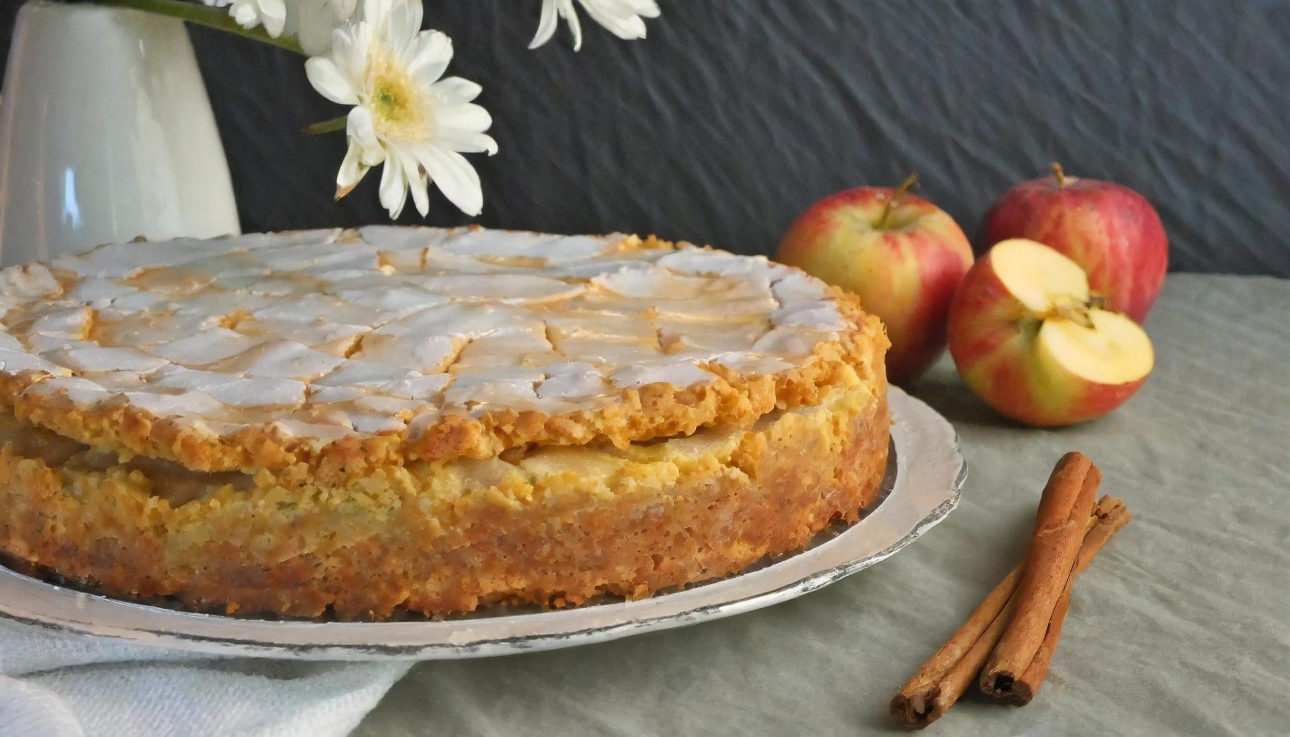 Ein frisch gebackener Apfelkuchen mit goldbrauner Kruste und glänzendem Zuckerguss auf einem silbernen Teller. Im Hintergrund stehen zwei ganze Äpfel und ein halbierter Apfel neben zwei Zimtstangen. Eine Vase mit weißen Blumen ergänzt das rustikale Arrangement auf einem weißen Leinentuch vor einer schwarzen, strukturierten Schieferwand.