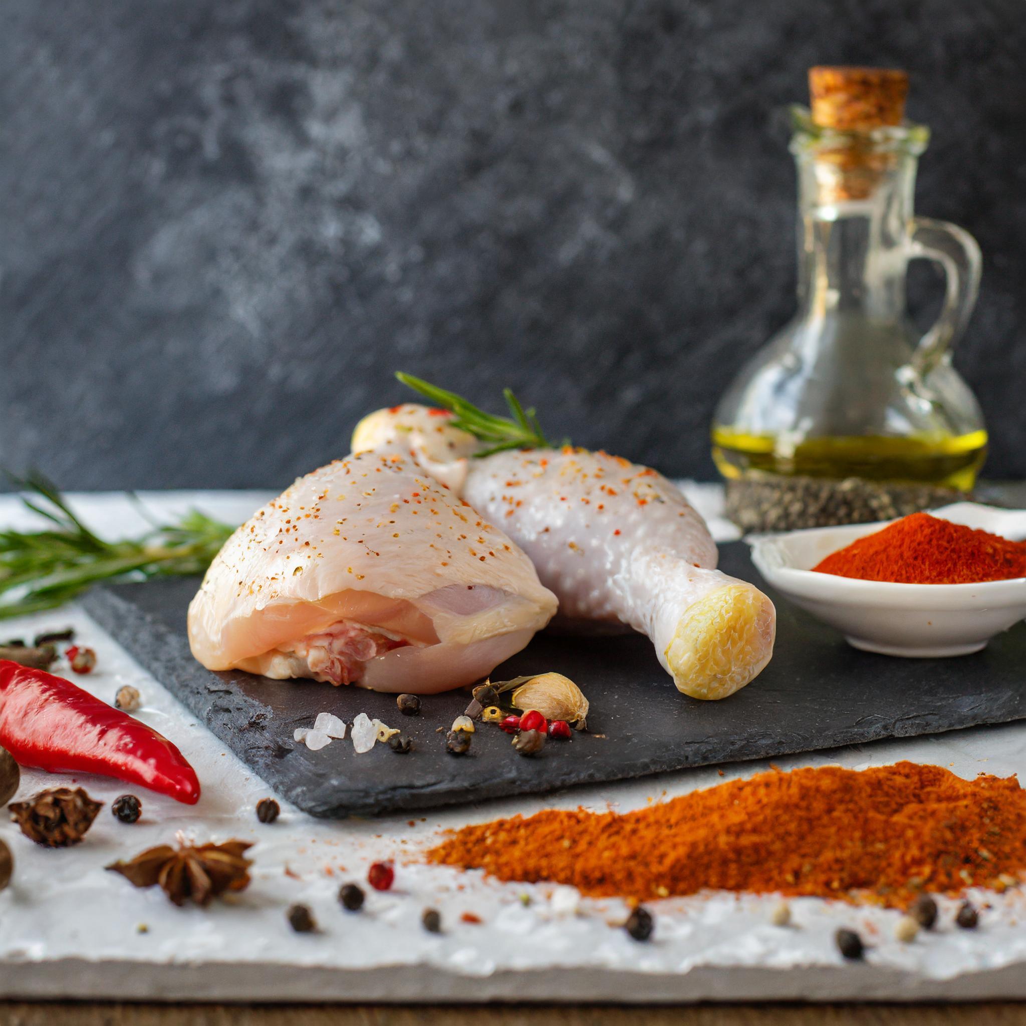 Ein Arrangement von rohem Hähnchen auf einem schwarzen Schieferbrett. Im Vordergrund sind eine gewürzte Hähnchenbrust und ein Hähnchenschenkel, garniert mit Rosmarin. Daneben liegen grobes Salz, Pfefferkörner, Paprikapulver und eine rote Chilischote. Im Hintergrund stehen eine Flasche Olivenöl und eine Schale mit Paprikapulver. Alles ist vor einem dunklen, strukturierten Hintergrund arrangiert.