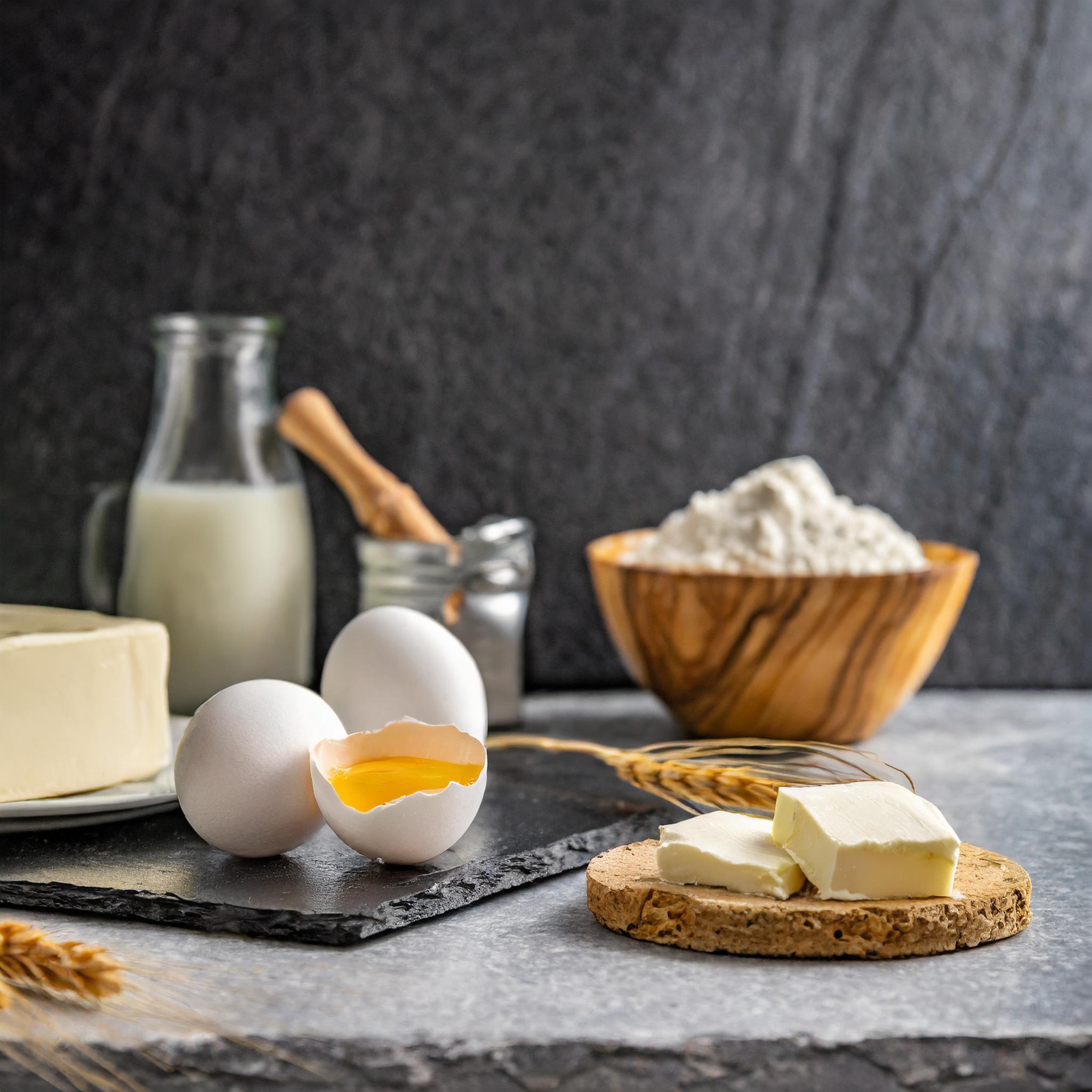 Eine rustikale Backszene mit zwei weißen Eiern, eines davon aufgeschlagen, auf einer schwarzen Schieferplatte. Daneben liegen ein paar Butterstücke auf einem runden Korkuntersetzer. Im Hintergrund stehen ein Krug Milch, ein Glas mit einem Holzlöffel, eine Holzschale mit Mehl und einige Weizenähren. Das Ganze spielt sich auf einer weißen Granitplatte vor einer dunkelgrauen, strukturierten Schieferwand ab.