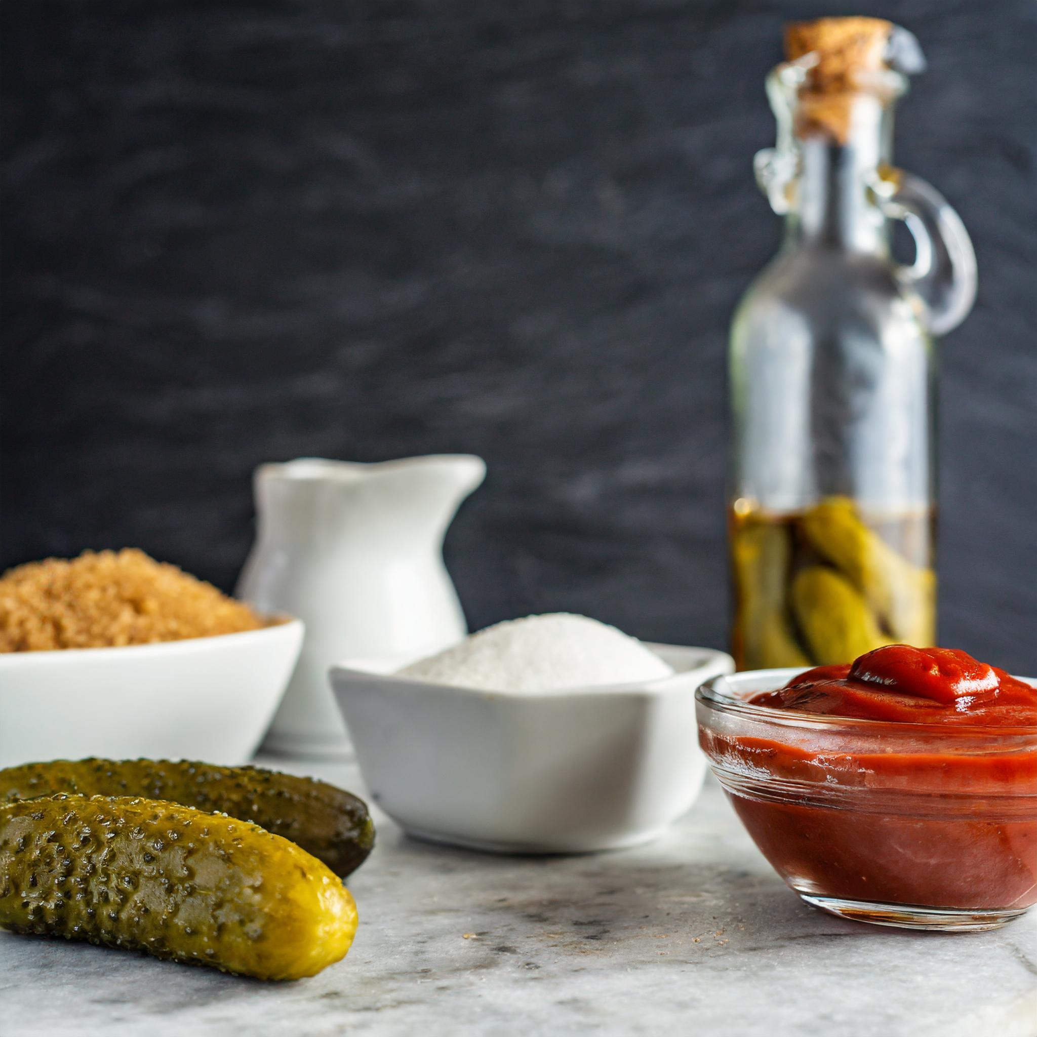 Ein rustikaler weißer Granittisch mit verschiedenen Zutaten: Im Vordergrund liegen zwei Essiggurken, daneben eine Schüssel mit rotem Ketchup. Im Hintergrund sind Schalen mit braunem und weißem Zucker sowie ein weißer Krug zu sehen. Eine Glasflasche mit Korkverschluss, gefüllt mit Essiggurken, steht ebenfalls im Hintergrund vor einem dunklen Schieferhintergrund. Die Szene wirkt einladend und bereitet den Eindruck einer leckeren Kochvorbereitung.