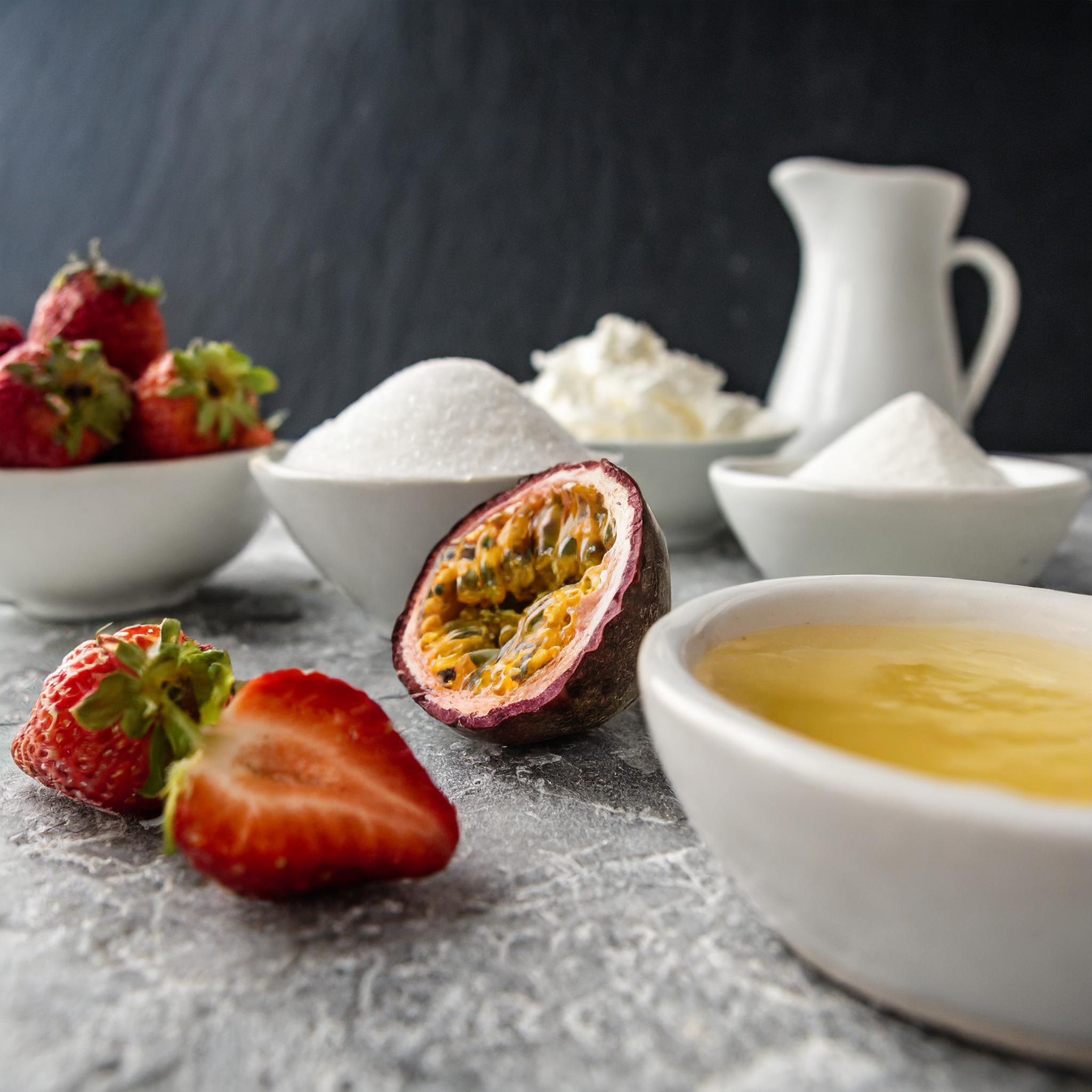 Eine Anordnung von Erdbeeren, Passionsfrucht, Zucker, Sahne und einer Schüssel mit gelbem Pudding auf einer grauen Oberfläche.