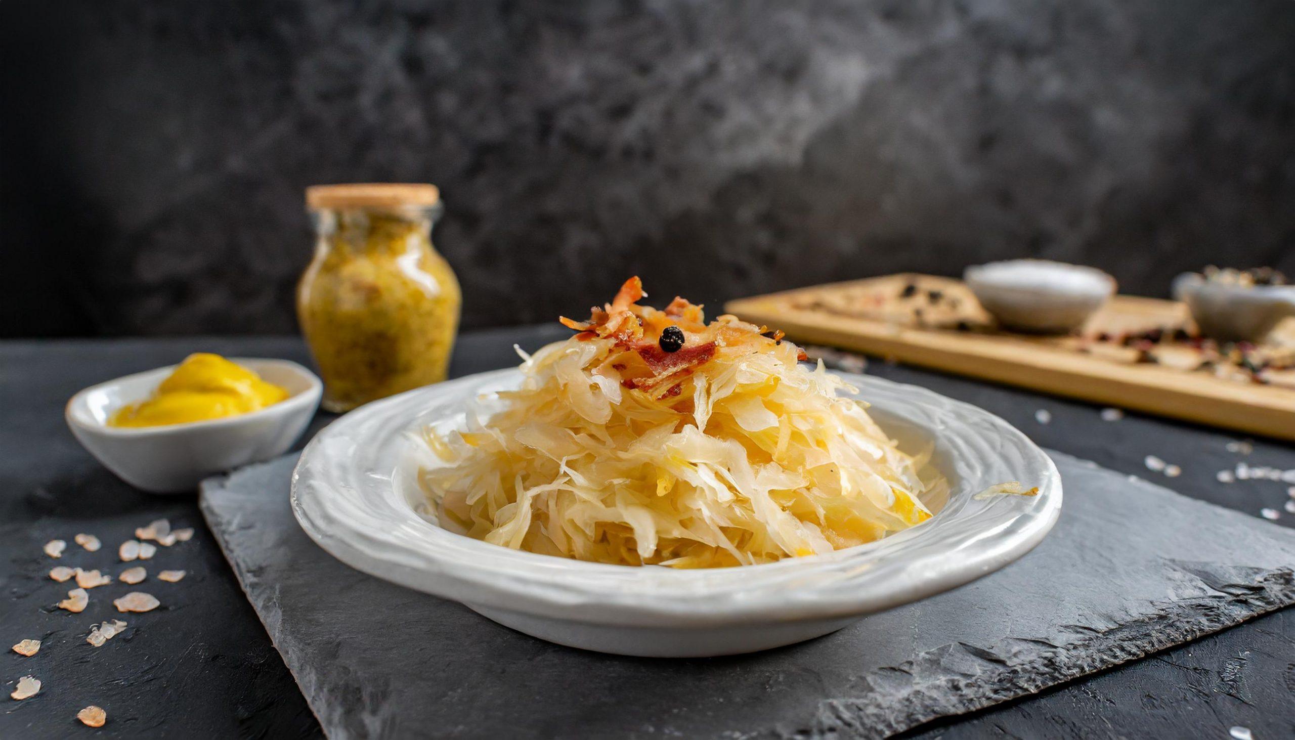 Ein Teller mit Sauerkraut, garniert mit Speckstückchen, neben Senf und einem Glas Senf im Hintergrund, auf einer dunklen Oberfläche.