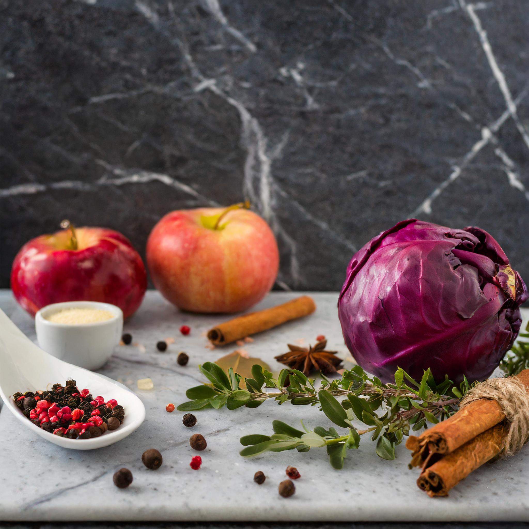 Ein Kopf Rotkohl, Äpfel, Zimtstangen, bunte Pfefferkörner und Zucker auf einem marmorierten Brett, umgeben von frischen Kräutern.