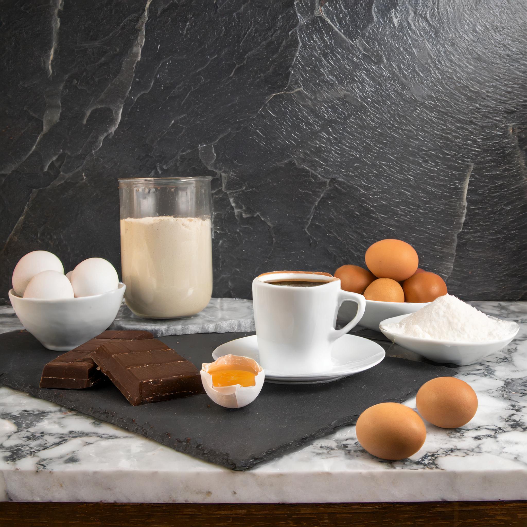 Ein Arrangement aus Eiern, Mehl, Zucker, Schokolade und einer Tasse Kaffee auf einem Marmor- und Schieferhintergrund.